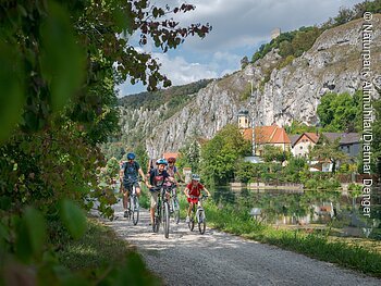 Gruppe von Radfahrern auf einem Schotterweg entlang eines Flusses, im Hintergrund Felsen und Gebäude.