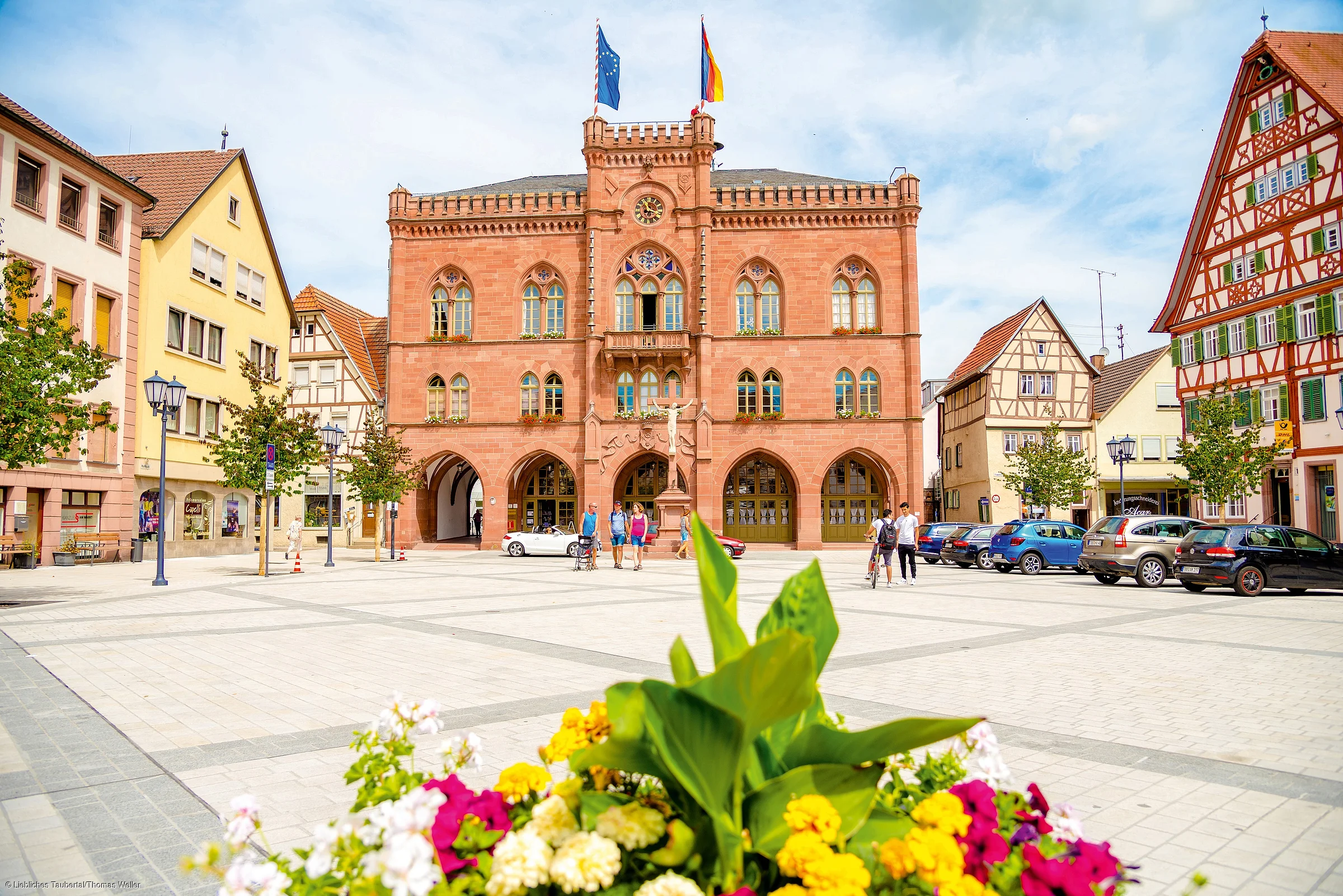 Ein Marktplatz mit einem roten Rathaus, davor Autos und Menschen