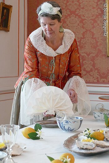 Frau in historischer Kleidung faltet eine weiße Serviette an einem gedeckten Tisch mit Zitronen und Muscheln.