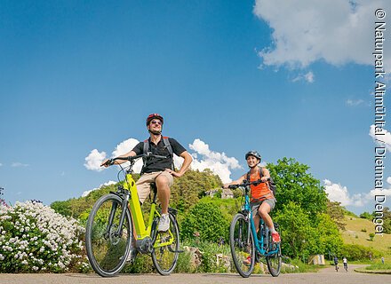Zwei Personen fahren bei sonnigem Wetter mit Fahrrädern auf einem Weg in grüner Landschaft.