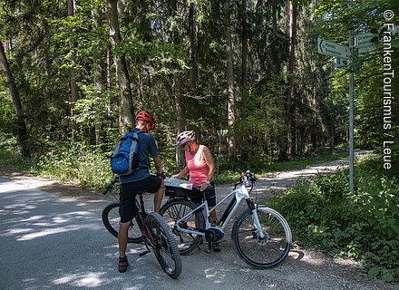 Zwei Radfahrer mit Helmen lesen eine Karte an einer Weggabelung im Wald bei Sonnenschein.