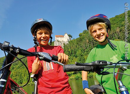 Zwei Jungen mit Fahrradhelmen und Fahrrädern vor bewaldetem Hügel und Schloss bei klarem Himmel.