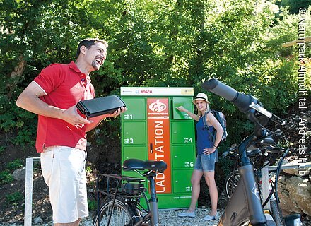 Mann hält Fahrradakku, Frau steht an grüner Ladestation für E-Bikes im Freien vor Büschen.