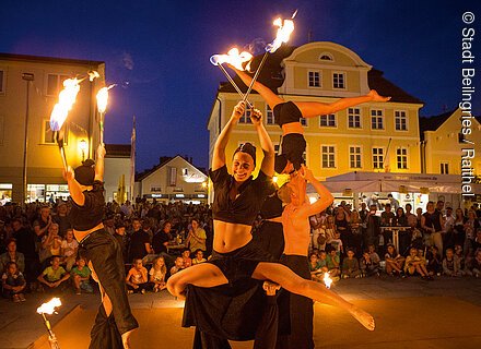 Feuershow mit vier Akrobaten und Publikum auf einem Platz vor beleuchteten Gebäuden bei Nacht.