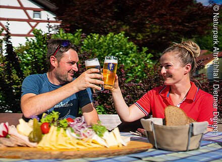 Zwei Personen stoßen mit Biergläsern an, davor eine Platte mit Käse, Wurst und Gemüse auf einem Tisch.