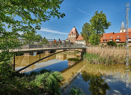 Fahrradfahrer auf einer Brücke über einen Fluss mit Bäumen und historischen Gebäuden im Hintergrund bei blauem Himmel.