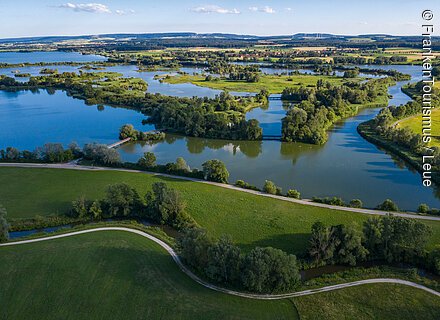 Landschaft mit Seen, Inseln, Bäumen und Wegen unter blauem Himmel, im Hintergrund Felder und Hügel.