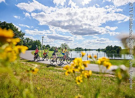 Tauber Altmühl Radweg - Rund um den Altmühlsee Vier Radfahrer fahren auf einem Weg am Flussufer bei bewölktem Himmel und blühenden gelben Blumen.