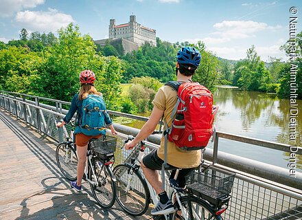 Radfahrer auf dem Tauber-Altmühl-Radweg (Eichstätt, Naturpark Altmühltal) Zwei Radfahrer mit Rucksäcken und Helmen auf einer Brücke über einen Fluss vor einer Burg auf einem bewaldeten Hügel.