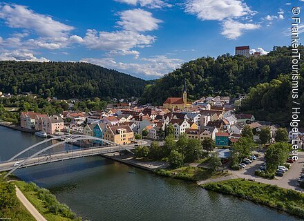 Riedenburg mit Rosenburg (Riedenburg, Naturpark Altmühltal) Blick auf eine Stadt mit Brücke über einen Fluss, umgeben von bewaldeten Hügeln und blauem Himmel.