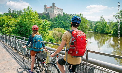 Zwei Radfahrer mit Rucksäcken und Helmen auf einer Brücke über einen Fluss vor einer Burg auf einem bewaldeten Hügel.