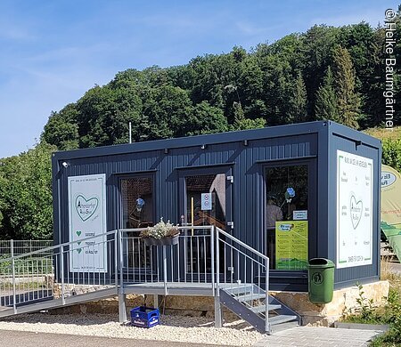 Kleines Verkaufsgebäude mit Rampe, umgeben von Grün und Wald im Hintergrund.