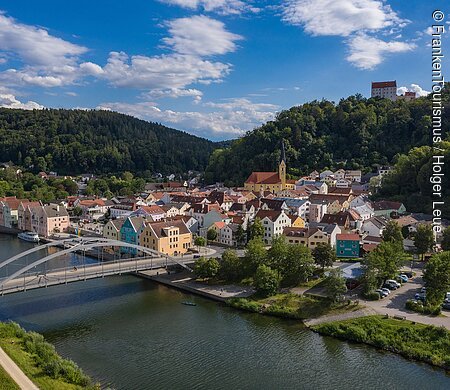Blick auf eine Stadt mit Brücke über einen Fluss, umgeben von bewaldeten Hügeln und blauem Himmel.