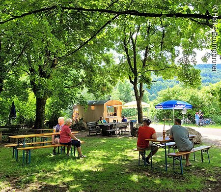 Menschen sitzen an Biergartenbänken unter Bäumen mit Imbisswagen und Sonnenschirmen im Hintergrund.