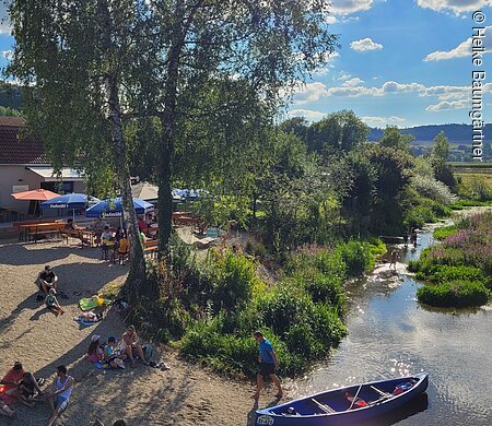 Menschen sitzen am Ufer eines Flusses mit Kanus und Bänken unter Bäumen an einem sonnigen Tag.