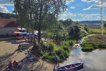 Menschen sitzen am Ufer eines Flusses mit Kanus und Bänken unter Bäumen an einem sonnigen Tag.