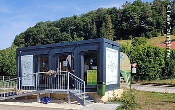 Kleines Verkaufsgebäude mit Rampe, umgeben von Grün und Wald im Hintergrund.
