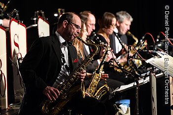 Fünf Musiker in schwarzer Kleidung spielen Saxophone in einer Reihe auf einer Bühne.