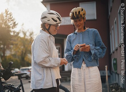 Radfahren auf dem Tauber Altmühl Radweg Zwei Personen mit Fahrradhelmen stehen draußen, eine zeigt der anderen etwas auf dem Smartphone.